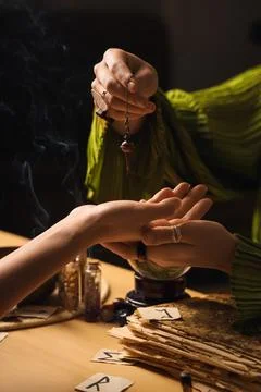 Fortune teller with pendulum reading woman's palm in room, closeup Stock Photos