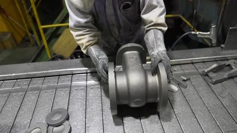 Foundry worker inspecting large cast iron valve body on roller conveyor after Stock Footage 336681597