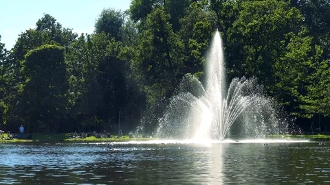 A fountain at a pond at a park on a sunny day, people at a shore and trees in Stock-Footage 73961053