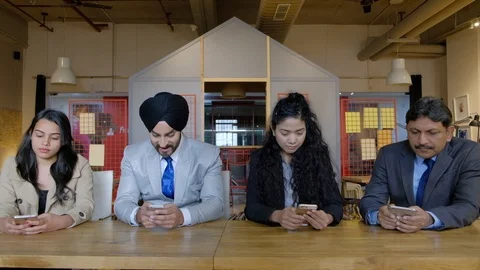 Four colleagues seated facing camera busy on the screen of their smart mobile ph Stock Footage 101889864