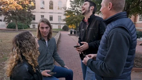 Four college students laughing while holding mobile phones sitting outside Stock Footage 104299315