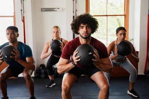 Four focused young adults doing medicine ball squats at the gym Stock Photos