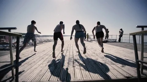 Four guys run down the pier and jump into the lake Stock Footage