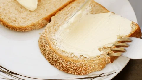 Four prong fork holds slice of bread as knife spreads white butter on top 스톡 동영상 128358016
