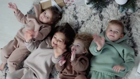 Four sweet children lay down jn a carpet near Christmas tree and waving their Stock Footage 145596673