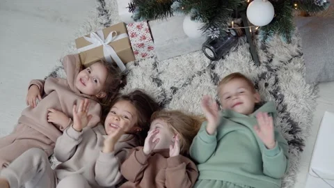 Four sweet children lay down jn a carpet near Christmas tree and waving their Stock Footage 145748067