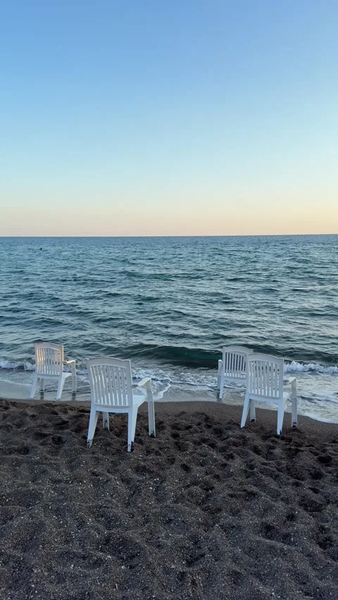 Four white plastic chairs placed on a sandy beach near the water's edge under Video stock 301847859