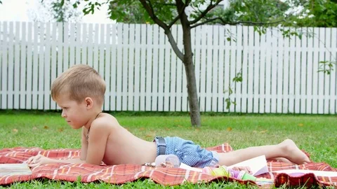 A four year old boy reads a book, in the garden, lying down on a blanket, a Stock Footage 80440140