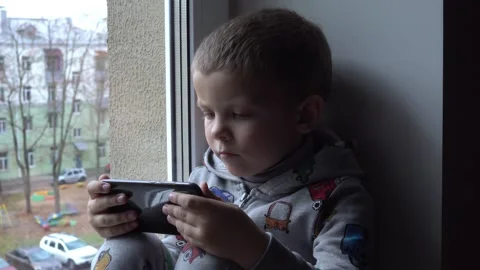 Four year old boy with smartphone on windowsill. View of autumn Park and street 스톡 동영상 142892441