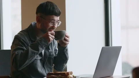 Freelancer drinking coffee while working in cafe. Smiling young man in glasses Stock Footage 234108967