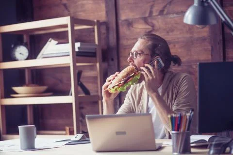 Freelancer eating sandwich while talking on phone. Stock Photos