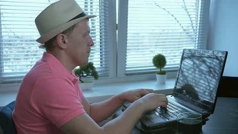 Freelancer in hat sitting at table and typing text on laptop, window background Stock Footage 155651595