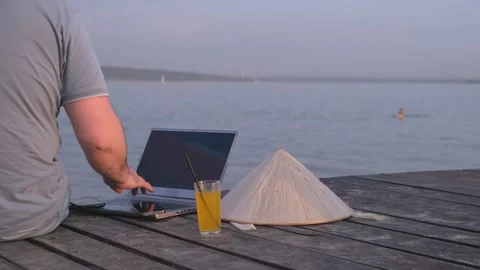 Freelancer man in hat holding smartphone, working on laptop computer by seashore Video stock 244485938