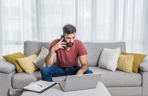 Freelancer man sitting on sofa talking on the phone and working on his laptop Stock Photos