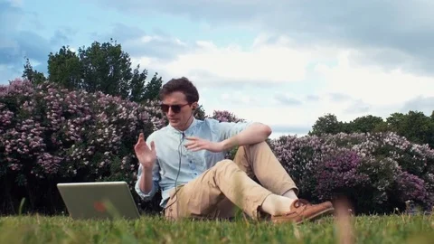 Freelancer talking on video calling with a laptop sitting on grass at city park Stock Footage 77168081