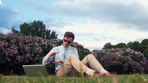 Freelancer talking on video calling with a laptop sitting on grass at city park Stock-Footage 77168117