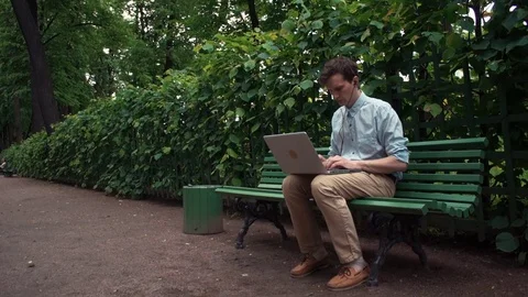 Freelancer using laptop with headphones sitting on bench. City park. Summer day Stock Footage 77168972