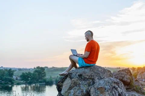 Freelancer working on a laptop while sitting on a cliff against the backgroun Stock Photos