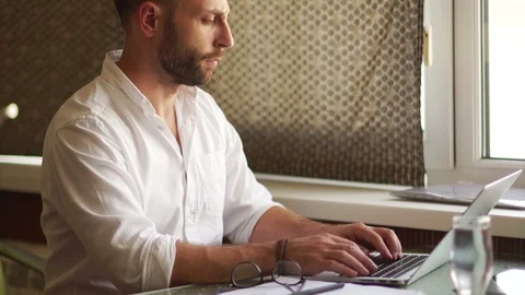 Freelancer working sitting at the table with a laptop. A young man in a white Stock Footage 97238359