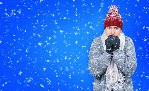 Freezing man in winter hat and scarf warming hands in falling snow on blue .. 스톡 사진