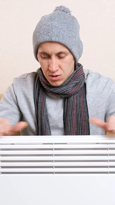 Freezing young man in warm clothes warming hands over an electric heater at home Stock Footage 344824221