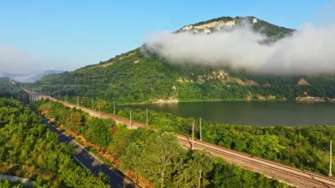 Freight train curving beside river through misty mountain valley with morning Stock Footage 341098664