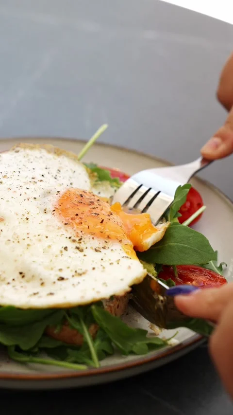 A fresh avocado toast with fried egg, arugula and cherry tomatoes is cut before Stock Footage 341401619