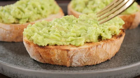 Fresh Avocado toast. Hands with fork spreading mashed avocado on bread Stock Footage 228691130