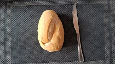 Fresh crusty bread roll served beside a sharp metal dinner knife Stock Photos