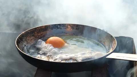 Fresh egg is fried in frying pan outdoors with oil and smoke on open fire Stock Footage 299805568