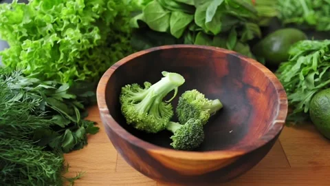 Fresh green broccoli florets fall into wooden bowl surrounded by lettuce Stock Footage 340026775