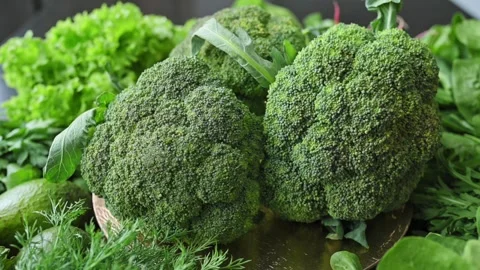 Fresh green broccoli rotates close up against backdrop of lettuce, spinach Stock Footage 340026612
