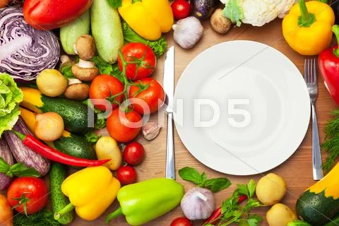 Fresh organic vegetables around white plate with knife and fork Stock Photos