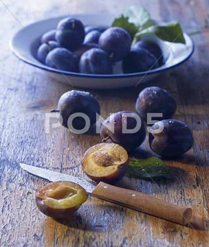 Fresh plums on a plate and on a wooden table, one halved Stock Photos