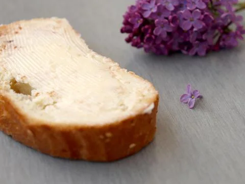 Fresh slice of bread with butter next to lilac flowers on gray surface during 스톡 사진