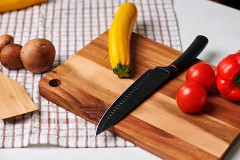 Fresh vegetables and a sharp knife arranged on a wooden cutting board with a che Stock Photos