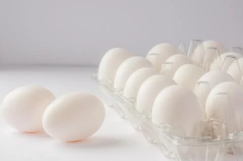 Fresh white eggs in a clear plastic carton with two eggs lying outside on a w Stock Photos