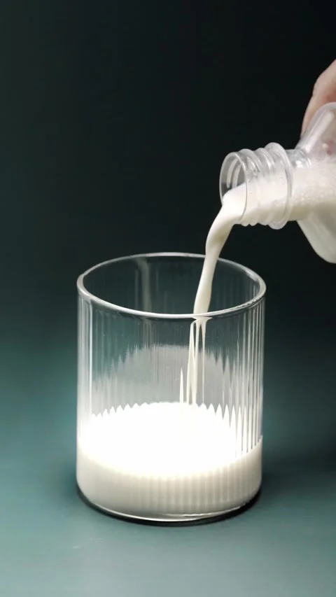Fresh white milk flows from a clear container into a vertically ribbed glas.. Видео 345125455
