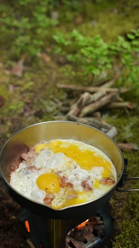 Fried eggs and bacon cooking in a stainless steel pan over a campfire stove Stock Footage 340653587