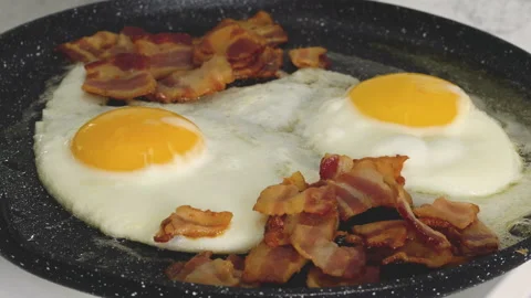 Fried eggs and crispy fried bacon slices close-up on a frying pan Stock Footage 238789763