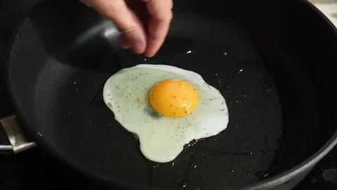 Fried eggs are cooked in frying pan. Female hands sprinkle salt on egg Stock Footage 340331620