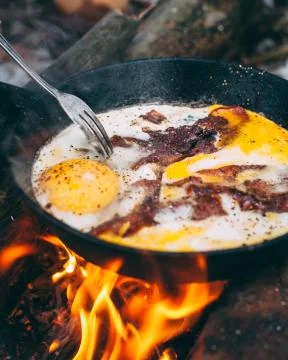 Fried eggs with bacon in a pan in the forest. Food at the camp. Fried egg wit Stock Photos