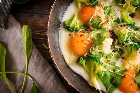 Fried eggs with cheese, broccoli, spinach in a pan on a dark wooden backgroun Stock Photos