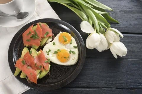 Fried eggs in heart shape and andwiches with avocado and smoked salmon on a dark Stock Photos