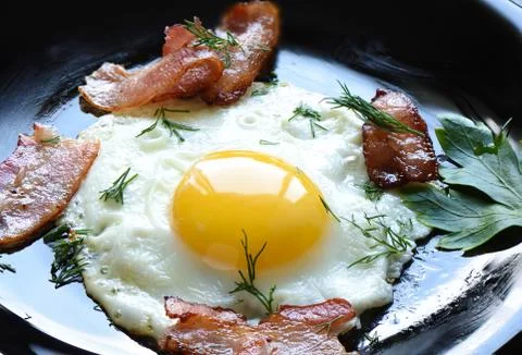 Fried eggs with slightly fried bacon and parsley on a balck plate. Stock Photos