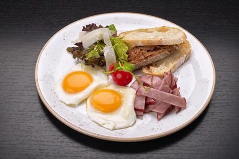 Fried fried eggs with tomatoes, ham and ciabatta toast, on a dark background. Stock Photos