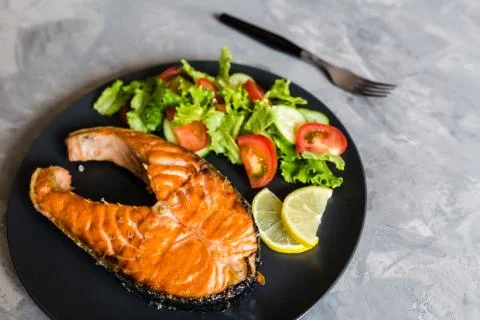 Fried salmon fish steak with vegetable salad and lemon on black plate. Creati Stock Photos