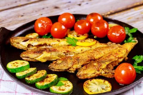 Fried small fish with tomato and lemon in a pan on a wooden background. Stock Photos