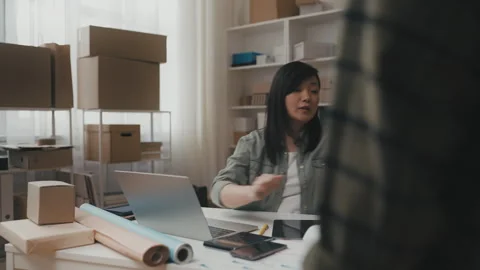 Friendly Asian post office worker giving parcel to customer, working laptop Stock Footage 251733754