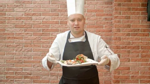 Friendly chef in uniform present a plate with seafood salad, talking to a camera 動画素材 74935886
